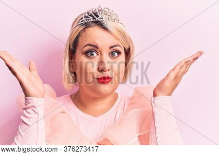 Beautiful blonde plus size woman wearing pincess tiara with diamonds over pink background clueless and confused with open arms, no idea and doubtful face.