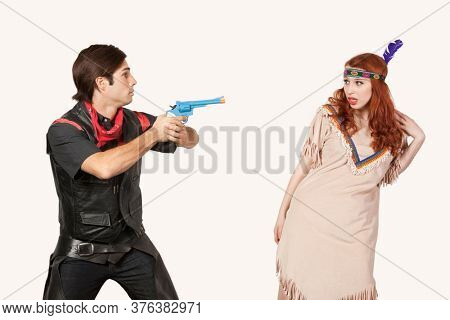 Young cowboy aiming gun at woman in old-fashioned costume against gray background