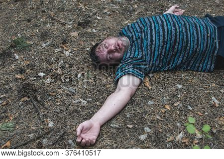 Murder In The Woods. The Body Of A Man In A Blue T-shirt Lies On The Ground Among The Trees In The F