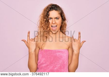 Beautiful blonde woman with blue eyes wearing towel shower after bath over pink background shouting with crazy expression doing rock symbol with hands up. Music star. Heavy concept.
