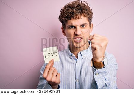 Young blond romantic man with curly hair holdiing paper with love message for valentines day annoyed and frustrated shouting with anger, crazy and yelling with raised hand, anger concept