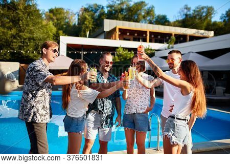Group Of Friends Having Fun At Poolside Summer Party, Clinking Glasses With Colorful Summer Cocktail