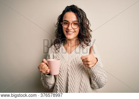 Young beautiful woman with curly hair drinking pink mug of coffee over white background happy with big smile doing ok sign, thumb up with fingers, excellent sign