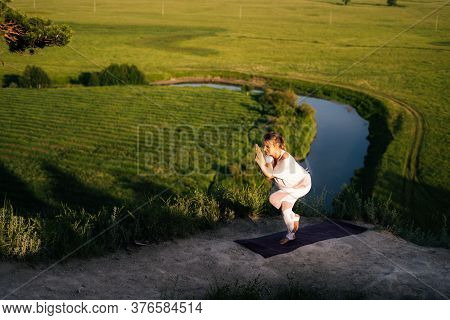 Young Woman Practicing Yoga, Standing In Eagle Exercise, Garudasana Pose On Top Of Cliff Background 