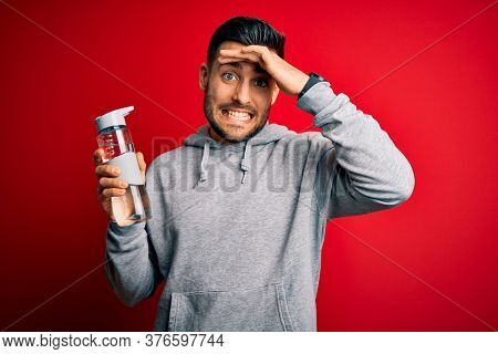 Young handsome sportsman drinking bottle of water to refeshment over red background stressed with hand on head, shocked with shame and surprise face, angry and frustrated. Fear and upset for mistake.