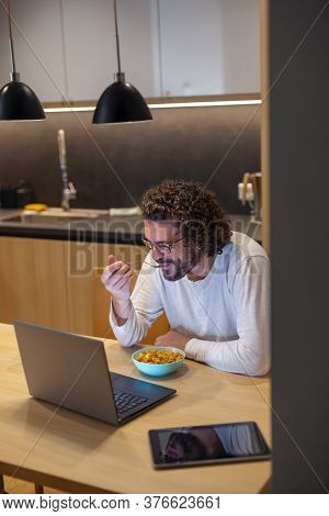 Handsome Young Man Sitting At Dinning Room Table, Wearing Pajamas And Eating Dinner While Watching A