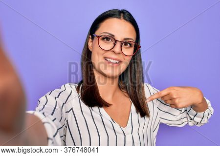 Young beautiful brunette woman wearing striped shirt and glasses making selfie by camera pointing finger to one self smiling happy and proud
