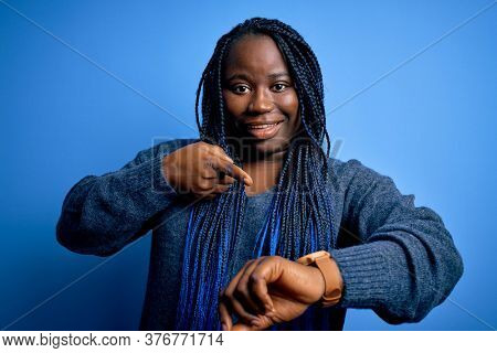 African american plus size woman with braids wearing casual sweater over blue background In hurry pointing to watch time, impatience, upset and angry for deadline delay