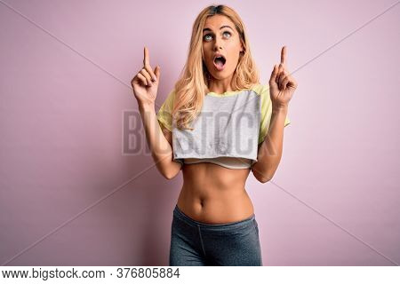 Young beautiful blonde sportswoman doing sport wearing sportswear over pink background amazed and surprised looking up and pointing with fingers and raised arms.