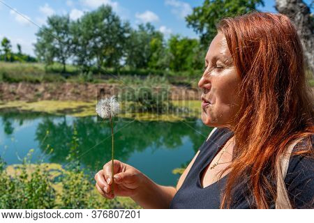 Beautiful Middle-age Woman With Red Hair Blowing A Dandelion In The Freshwater Pond Of Clot De La Ma