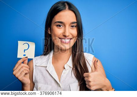 Young beautiful brunette woman holding paper with question mark symbol message happy with big smile doing ok sign, thumb up with fingers, excellent sign