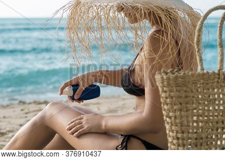 Beautiful Young Woman On The Beach Applying Sunblack While Sitting On Her Towel And Wearing Big Stra