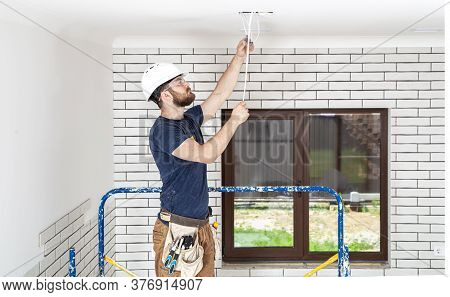 Electrician Builder At Work, Installation Of Lamps At Height. Professional In Overalls With An Elect