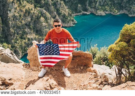 Young Man Sitting On A Mountain Top And Holding The Us Flag In His Hands. Veteran Traveller Holding 