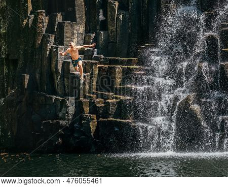 Teenage Boy Jumping In Waterfall Lake. Falling Water Streams Flow On Black Volcanic Stone Cascades. 