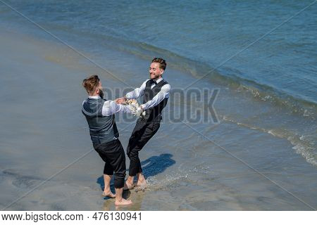 Gay Grooms Walking Together On Sea Beach During Wedding Day. Romantic Men In Sea Water. Homosexual C