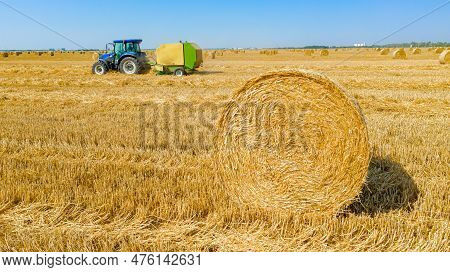 Above View Round Bale Of Straw After Harvest, In Background Tractor Is Pulling Round Baler, Machine 