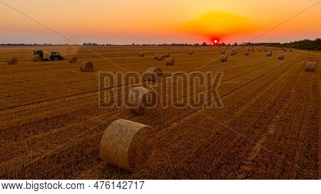 Above View Round Bales Of Straw After Harvest, In Background Tractor Is Pulling Round Baler, Machine