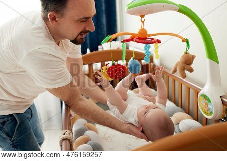 A Happy Dad Puts His Baby In A Crib With A Musical Toy. The Father Hugs His Baby. Happy Childhood.