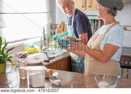 Cheerful Senior Couple In The Home Kitchen While Wife Holds A Freshly Baked Homemade Plumcake And Hu