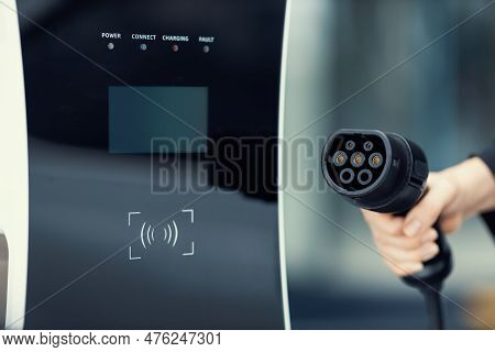 Focus Closeup Hand Holding Ev Plug For Electric Vehicle Pointing At Camera With Charging Station At 