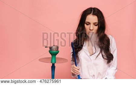 Young, Attractive Woman Smokes A Hookah, Sheesha On A Pink Background. The Pleasure Of Smoking.