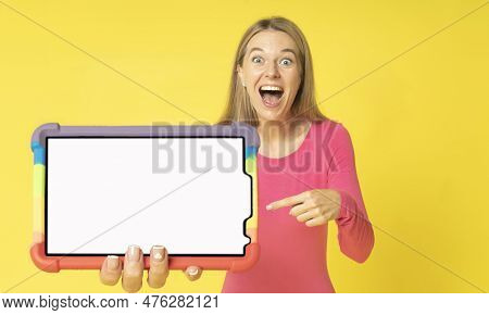 Excited young woman pointing to tablet computer with white mock-up screen in hand showing or presenting isolated on yellow background. Attractive amaized woman holding big tablet pc in rainbow cover