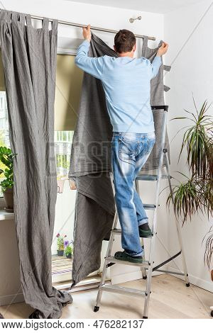 A Man Stands On A Stepladder And Hangs Gray Curtains On A Curtain Rod In An Apartment, Spring Cleani