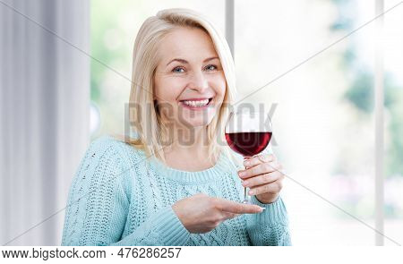 Happy Woman Holds A Glass Of Red Wine And Looks Relaxed At The Camera Sitting Near The Window.