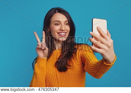 Cheerful Pretty Girl Dressed In Warm Knitted Orange Sweater Showing Vsign Gesture While Taking Selfi
