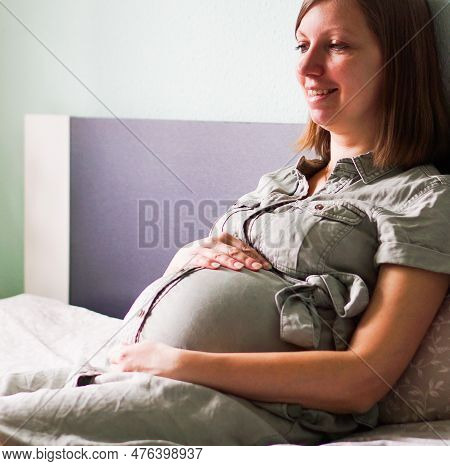 Happy Smiling Pregnant Woman Sitting On A Bed In A Bedroom At Home And Hugging Her Belly.