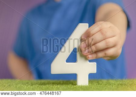 Number 4 In Hand. Hand Holding White Number Four Over Green Grass And Purple Background, Birthday Co