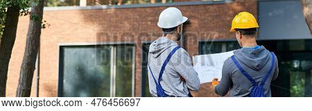 Making The Best House. Rear View Of Two Builders Standing Outdoors With An Open Blueprint Discussing
