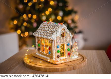 A Gingerbread House Against A Decorated New Years Eve Room With Lights