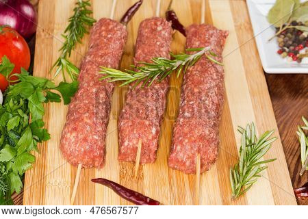 Several Raw Lula Kebab Skewered On Wooden Skewers On The Wooden Cutting Board Among The Some Vegetab
