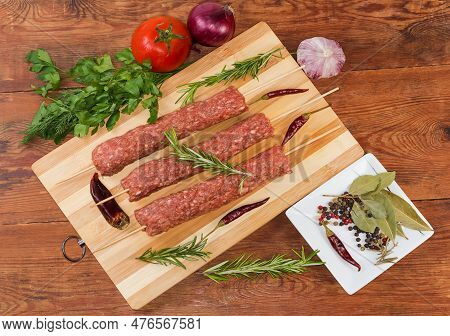 Several Raw Lula Kebab Skewered On Wooden Skewers On The Wooden Cutting Board Among The Some Vegetab