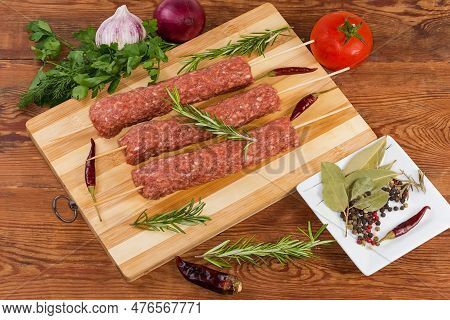 Several Raw Lula Kebab Skewered On Wooden Skewers On The Wooden Cutting Board Among The Some Vegetab