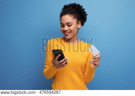 Young Latin Woman With Afro Hair Pays For A Purchase With A Phone Using A Card On A Studio Backgroun