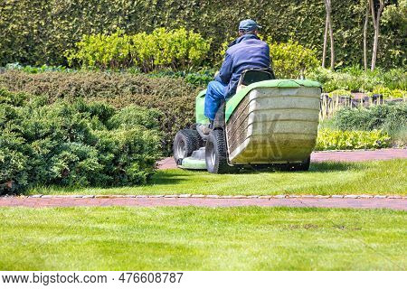 A Working Gardener Drives A Tractor Lawn Mower And Mows The Green Grass Of The Lawn Against A Blurre