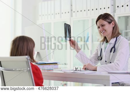 Female Pediatrician With Smile Holds X-ray Of Little Patient Sitting At Table At Workspace. Schoolgi