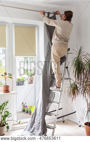 A Woman Stands On A Stepladder And Hangs Gray Curtains On A Curtain Rod In An Apartment, Spring Clea