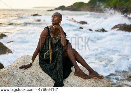 Androgynous Ethnic Fashion Model In Luxury Dress, Jewelry Sits On Rocks By Ocean. Gay Black Person I