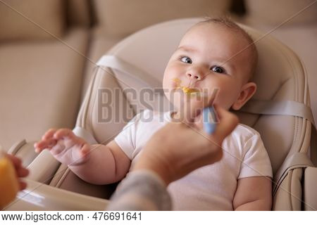 Mother Feeding Cute Baby Boy With Porridge, Baby Sitting In High Chair All Messy And Staied