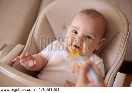 Mother Feeding Cheerful Baby Boy With Porridge, Baby Sitting In High Chair All Messy And Staied Smil