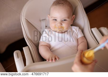 High Angle View Of Mother Feeding Cheerful Baby Boy With Porridge, Baby Sitting In High Chair All Me