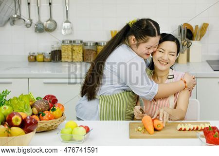 Asian Young Daughter Wears Apron Hugging Cuddling Showing Love To Mother Sitting Cutting Sweet Peppe