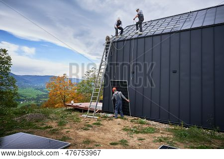 Men Mounters Installing Solar Panel System On Roof Of House. Workers In Helmets Lifting Up Photovolt