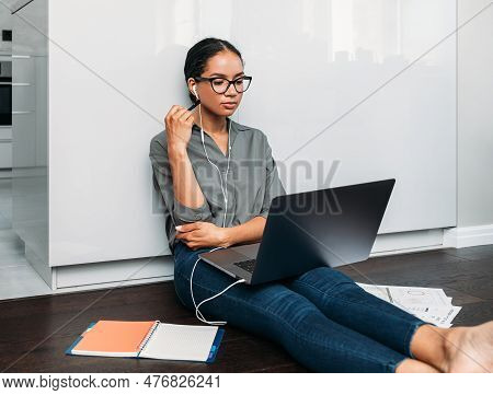 Young Female Entrepreneur In Thought Sitting On The Floor In The Kitchen With A Laptop On Her Legs