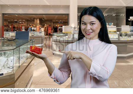 Portrait Of A Brunette Asian Woman Advertising A Red Gift Box. Boutique Mall Interiror In The Backgr