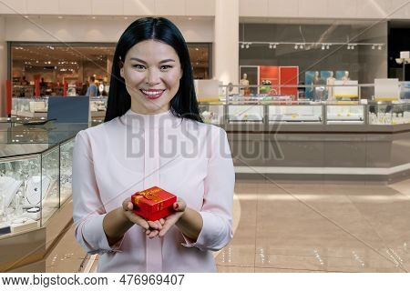 Smiling Brunette Asian Woman Giving You A Red Gift Box With Both Hands. Boutique Mall Interiror In T